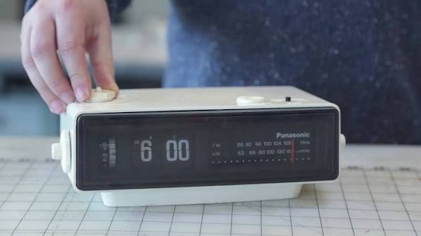 A hand on a white flip-clock radio showing 6:00.