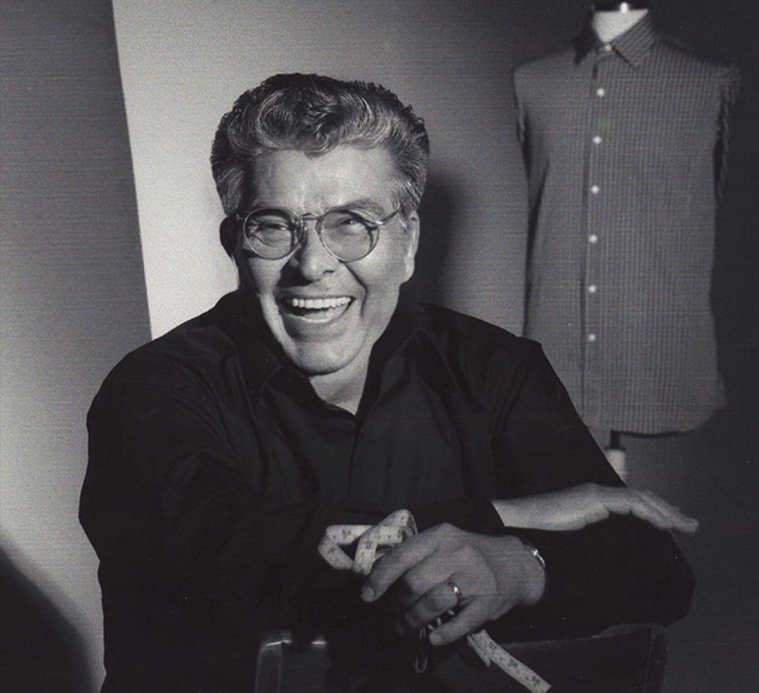 Ike Behar laughing, in glasses, holding a tape measure, a shirt on a dress form behind him.