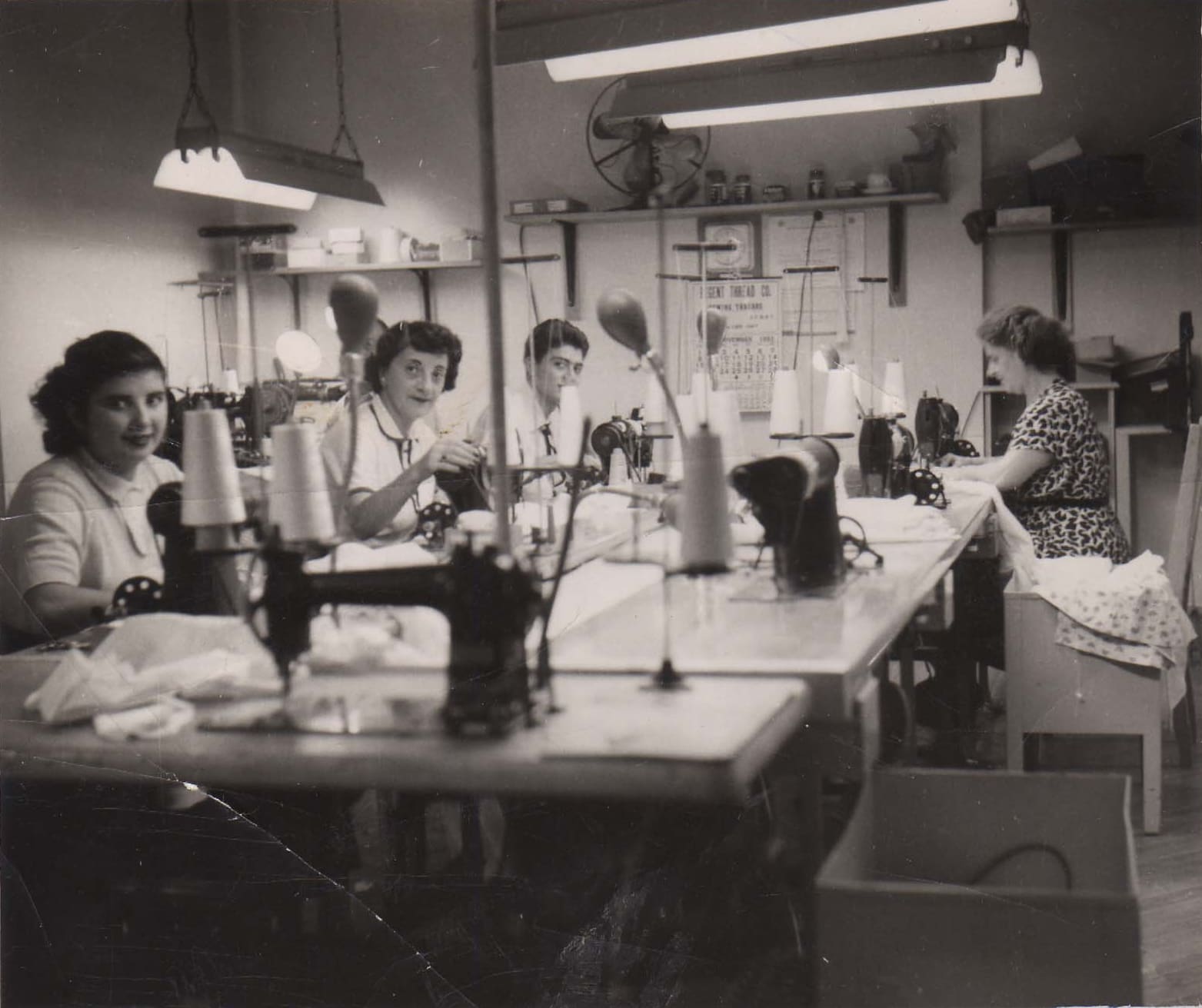 Four women at sewing machines in a small shirt workroom, in black and white.