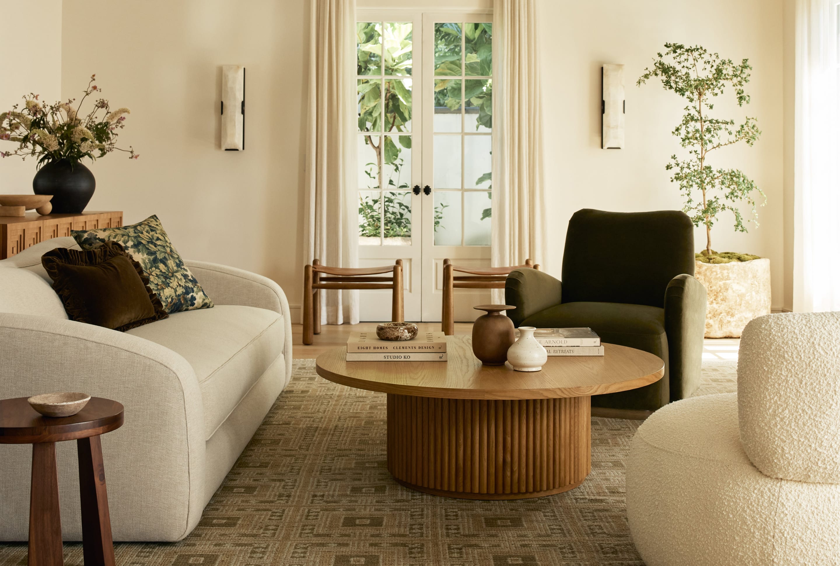 A bright living room: a curved cream sofa, a boucle chair, a green armchair and a fluted round wood coffee table