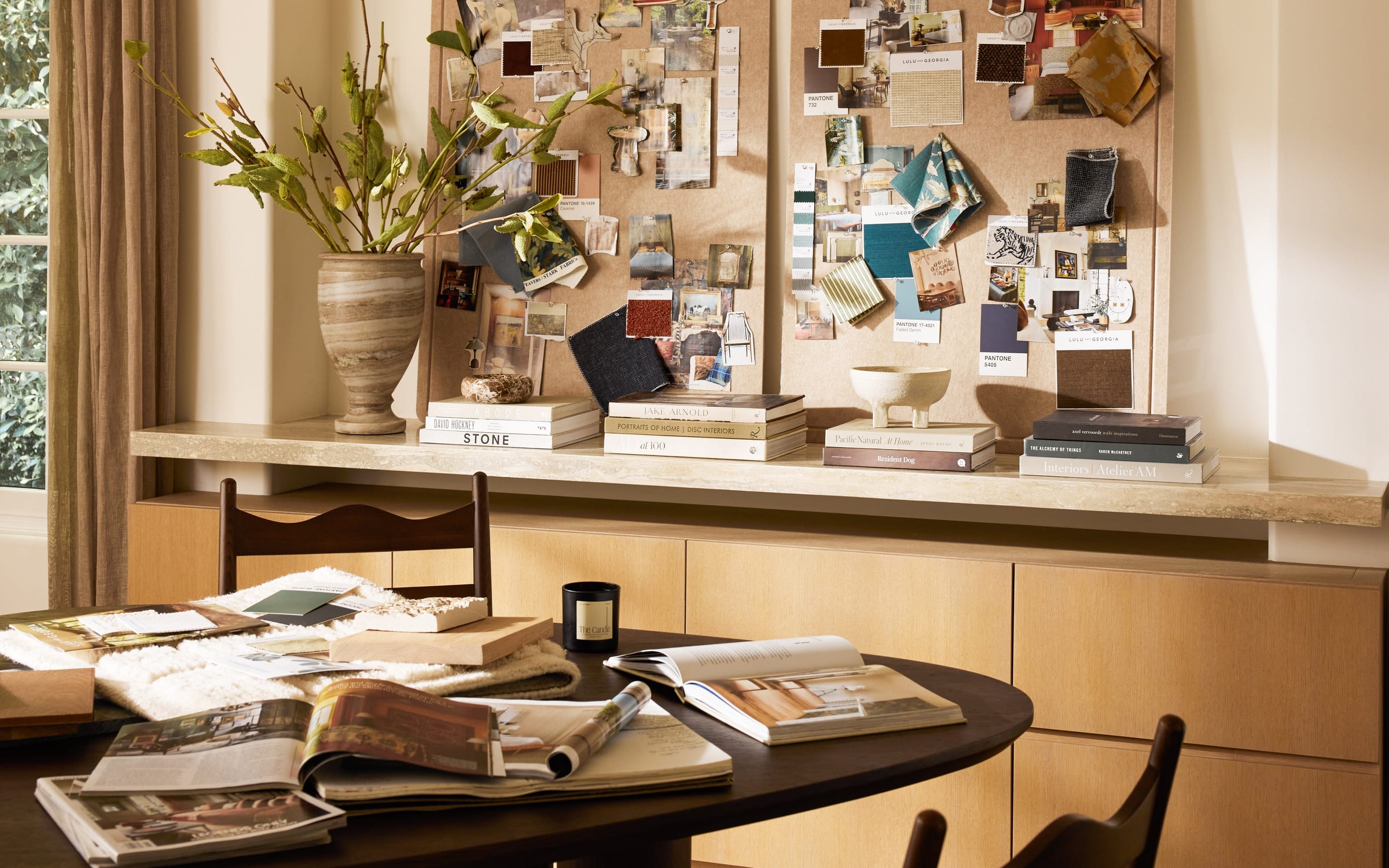 A styled work corner: pinboards of swatches and tear sheets, an oval table with magazines and a sample book.