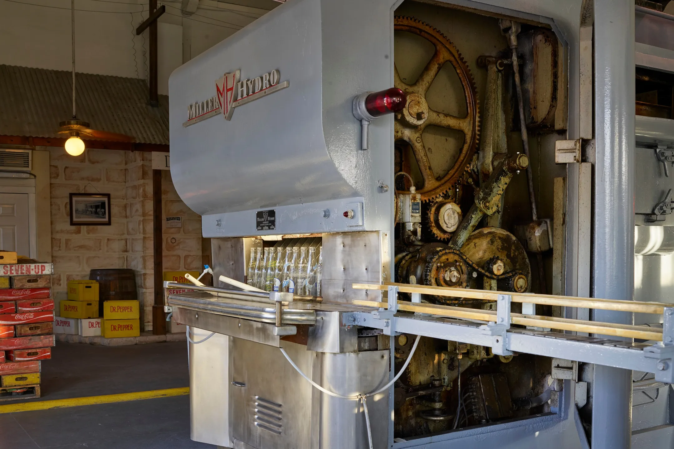 A vintage bottle-filling machine with a large gear wheel, a conveyor of bottles running beneath it.