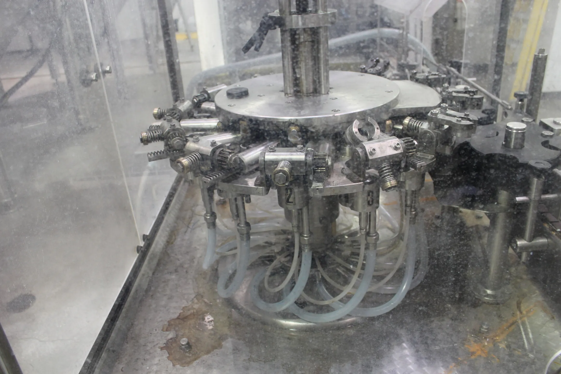 The filling head of a bottling machine behind glass, steel tubes curving down from a ring of valves.
