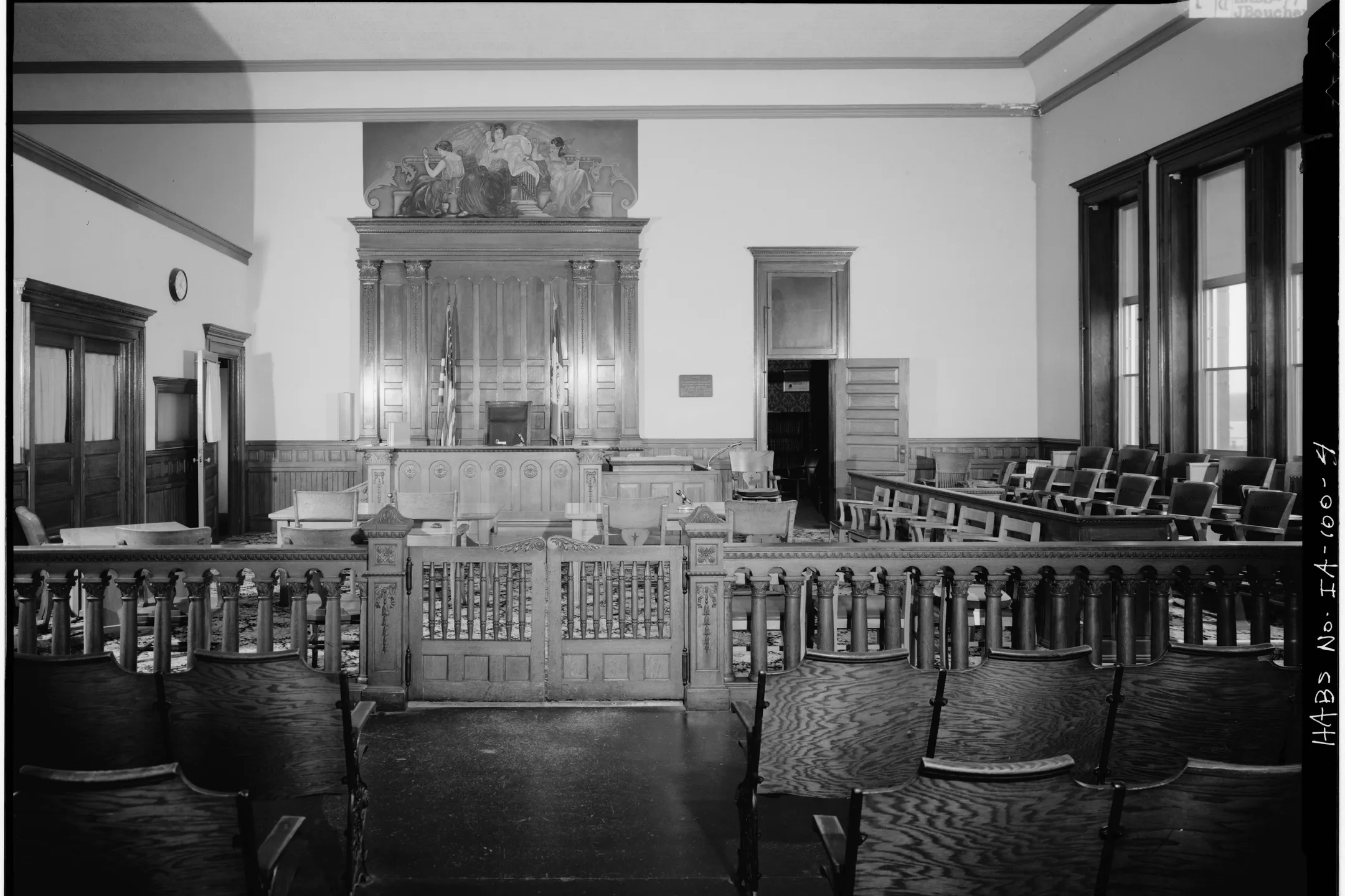 A historic courtroom in black and white: a rail and rows of seats before the judge’s bench.