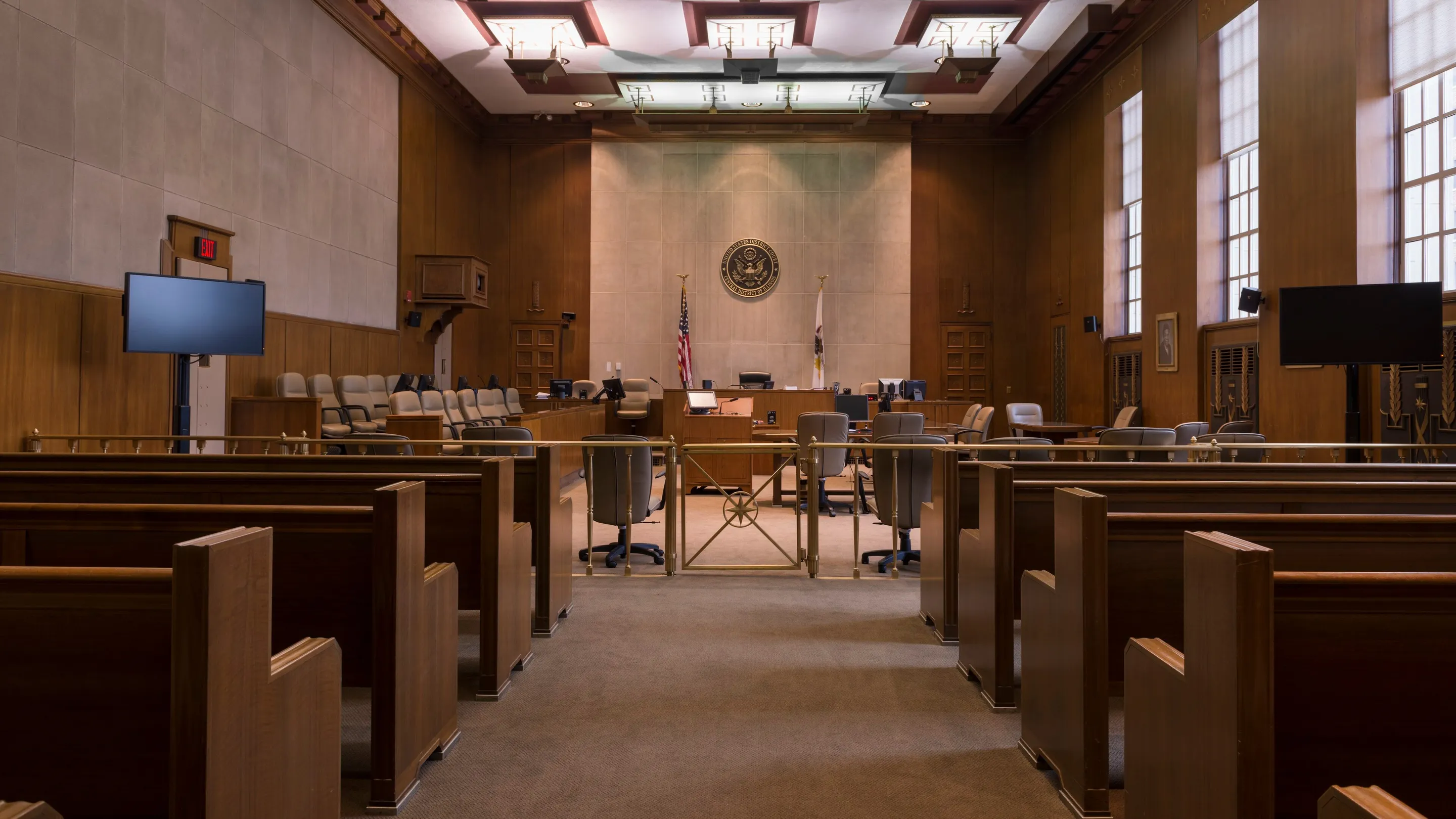 An empty wood-panelled courtroom: rows of pews, the well and the bench beneath a round seal.