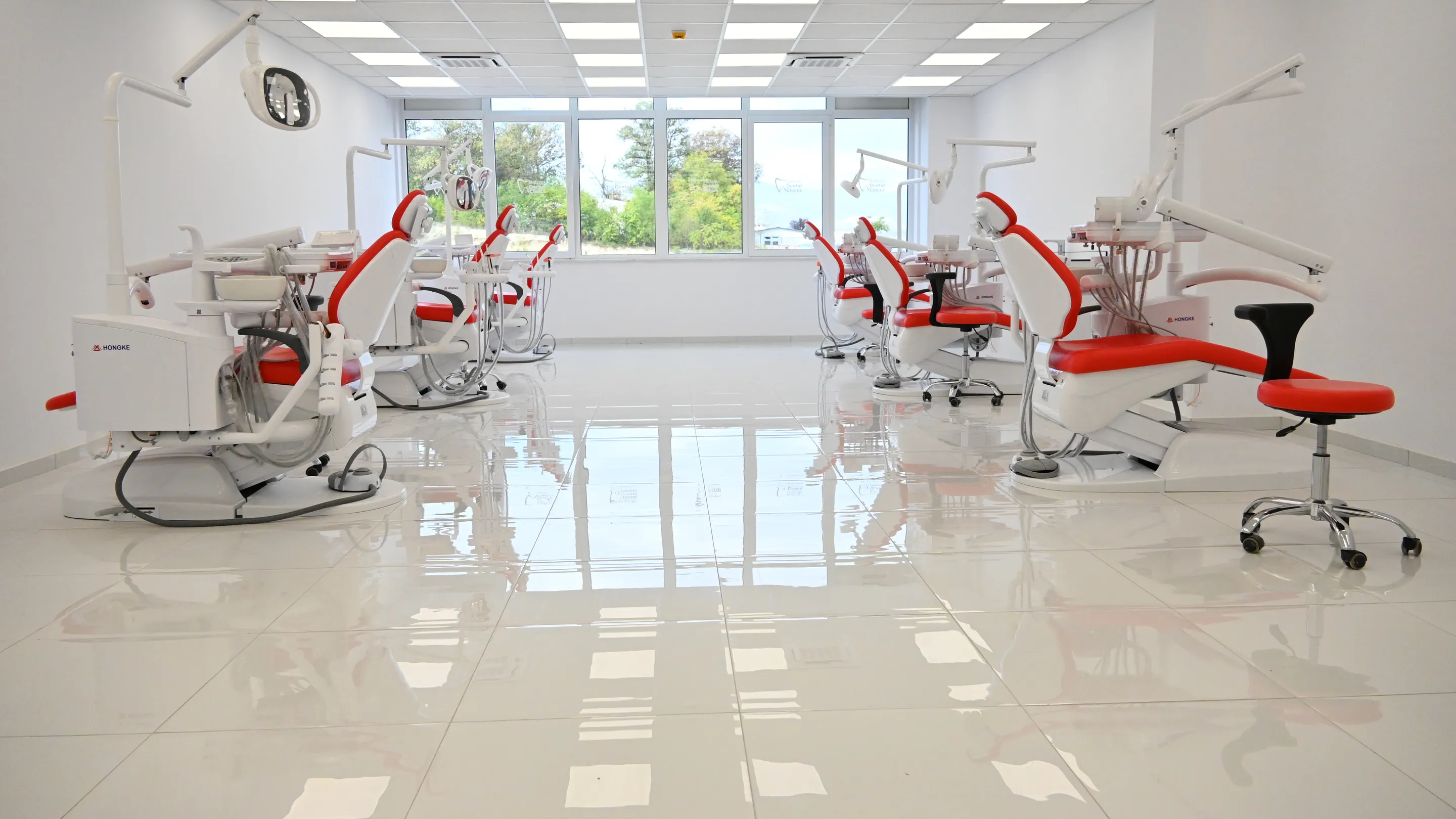 A bright open clinic: rows of dental chairs with red upholstery under ceiling lights, a wall of windows at the far end.