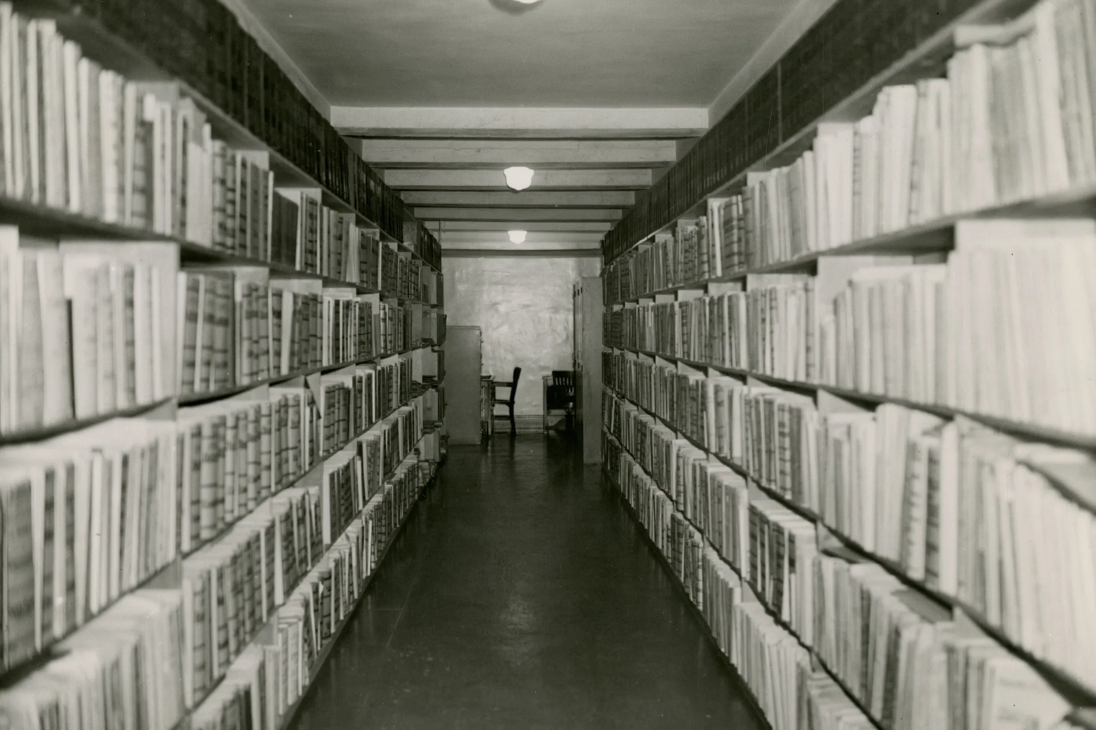A long aisle of library stacks in black and white, shelves of bound volumes on both sides.
